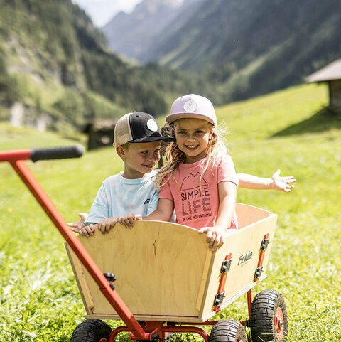 Ein Mann zieht zwei lachende Kinder in einem klassischen Holzwagen