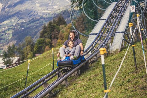 Ein Mann und ein Mädchen fahren mit einer Sommerrodelbahn.