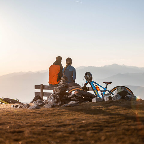 Zwei Radfahrer machen eine Pause auf einer Parkbank und blicken auf eine traumhafte Bergkulisse.