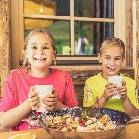 Almen & Hutten Ein Junge und ein Mädchen sitzen vor einem Kaiserschmarrn und trinken ein Glas Milch